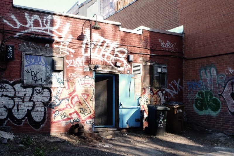 A back alley of a building with graffiti and a waste disposal bin.