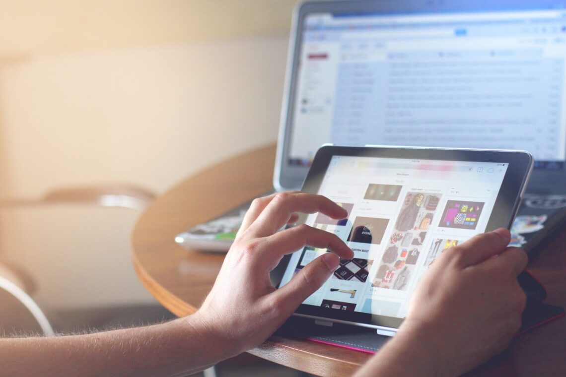 A pair of hands holds up a tablet and scrolls through an online store with a laptop behind them, displaying the changes in user behaviour that Google Analytics is trying to address with GA4.